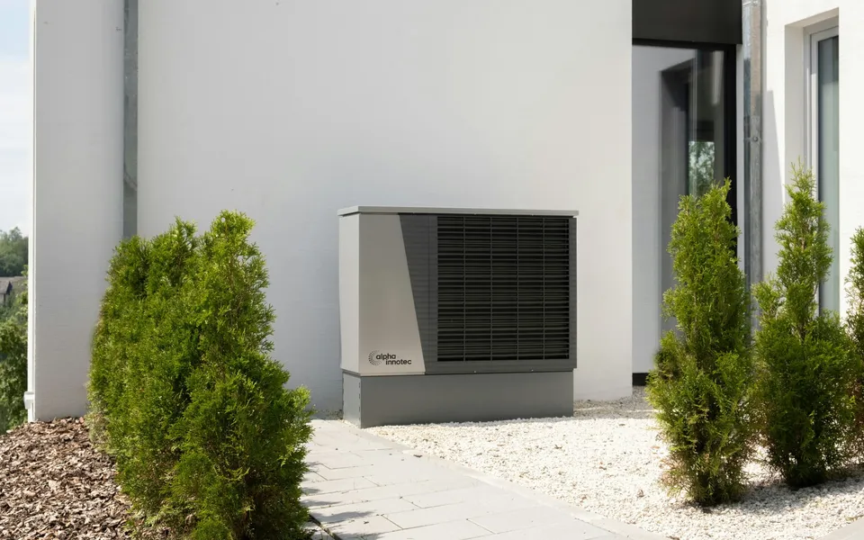 Außeneinheit einer Wärmepumpe neben einem weißen Wohnhaus mit Kieselbett und Zypressen